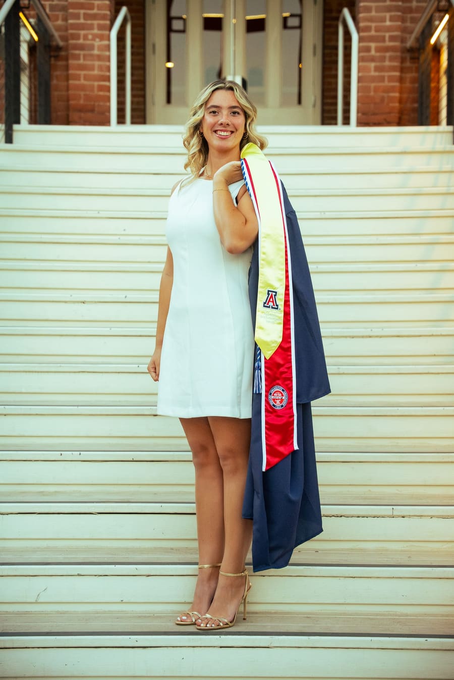 Graduate in a white dress holding her gown and stole on sunlit stone steps