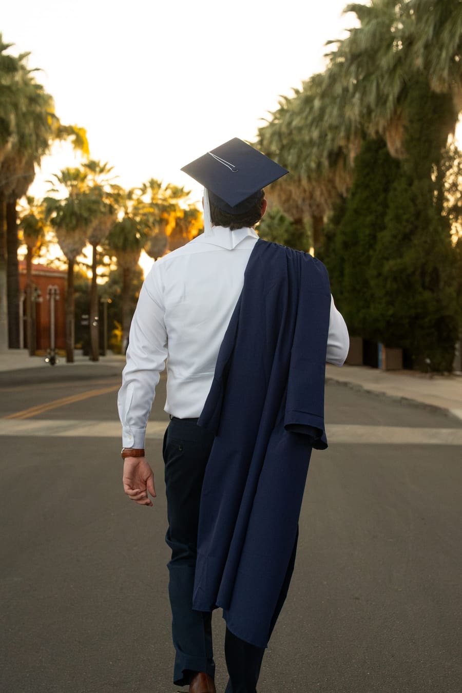Graduate walking away down a palm-lined street, gown over his shoulder